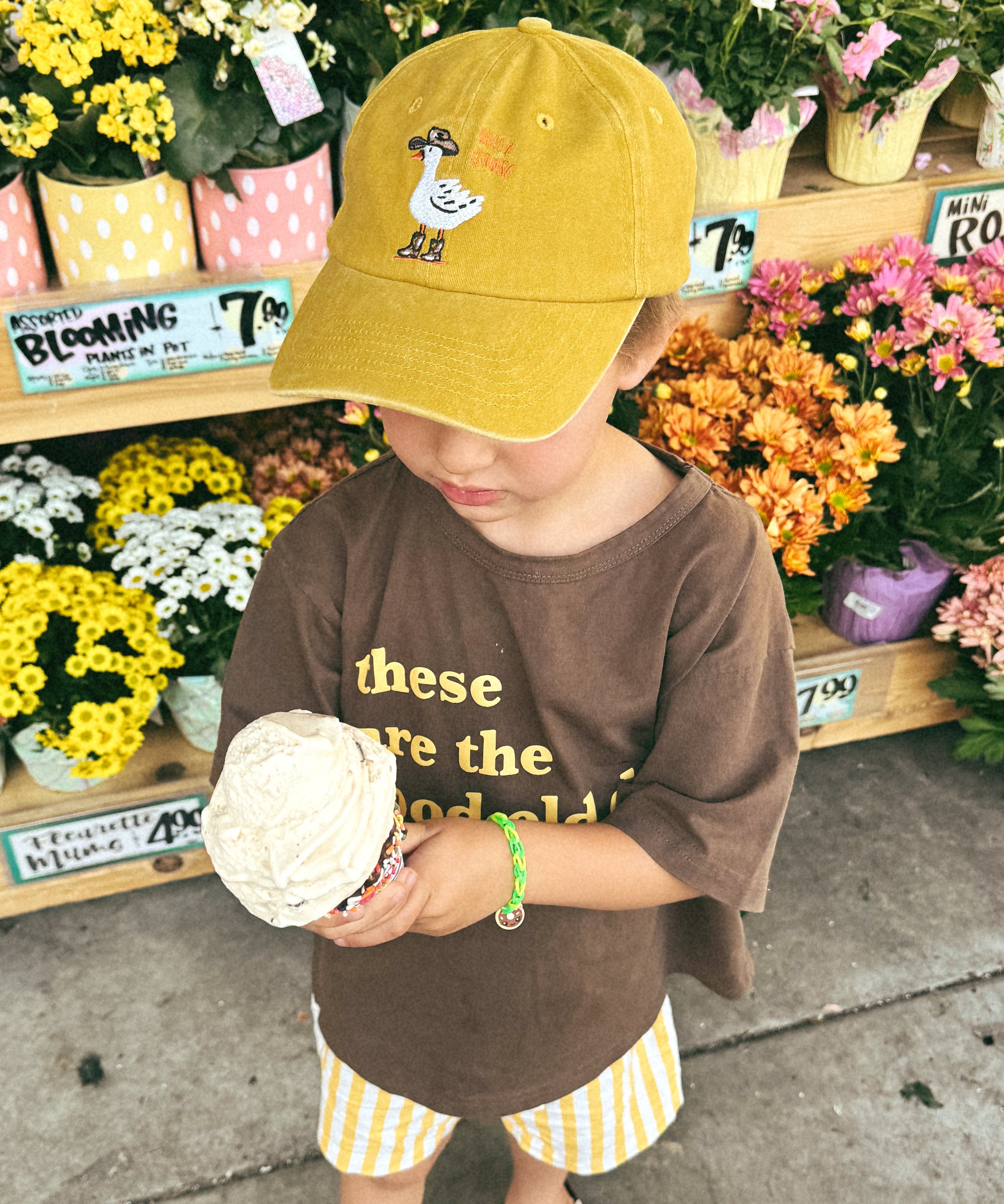 Person standing in front of flower arrangement with a yellow cap and brown shirt while holding an ice cream cone.