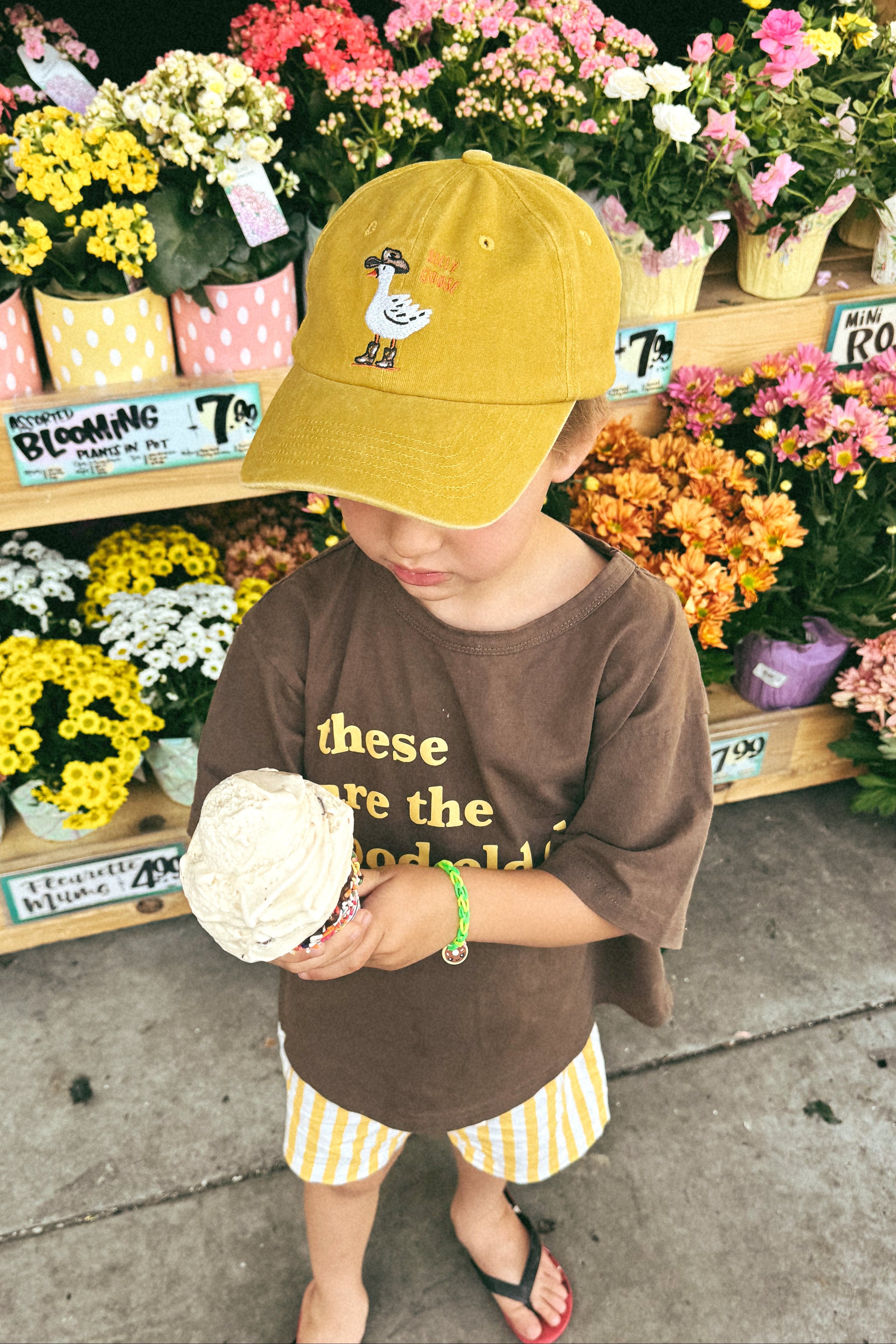 Person standing in front of flower arrangement with a yellow cap and brown shirt while holding an ice cream cone.
